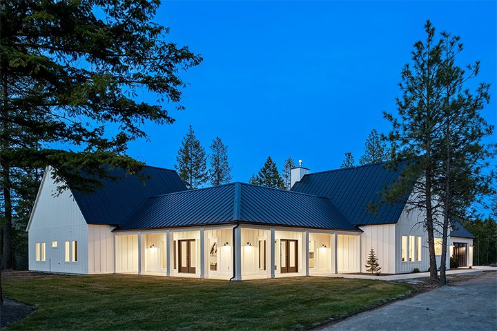Modern farmhouse-style home at dusk featuring board and batten siding, expansive windows, and a black metal roof. This High-End Residential Remodel by Schaff Homes Design-Build Contractor Spokane WA blends timeless architecture with contemporary design. Surrounded by evergreens, the illuminated wraparound porch highlights the craftsmanship and thoughtful design tailored for luxury living in the Pacific Northwest.